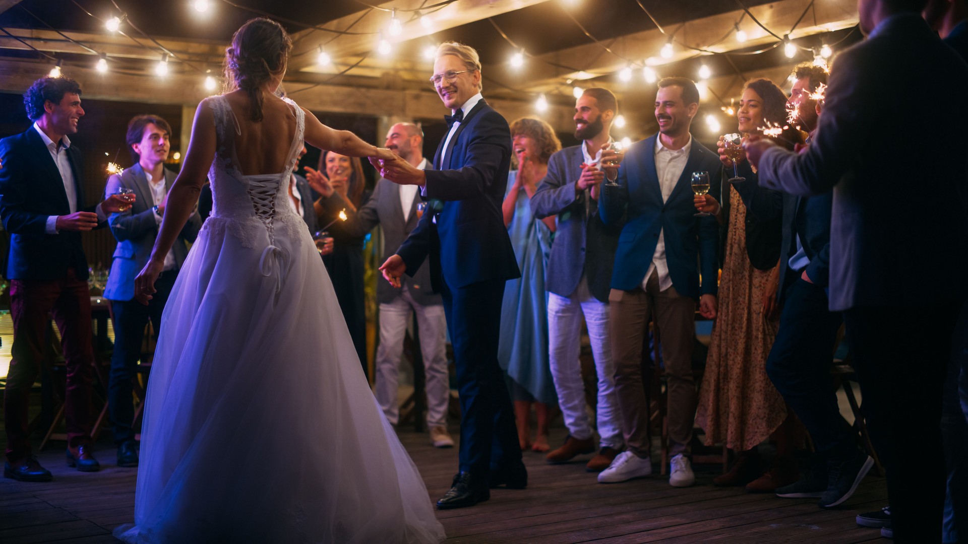 Beautiful Bride in White Dress and Groom in Stylish Black Suit Celebrate Wedding at an Evening Reception Party. Newlyweds Dancing at a Venue with Best Multiethnic Diverse Friends.